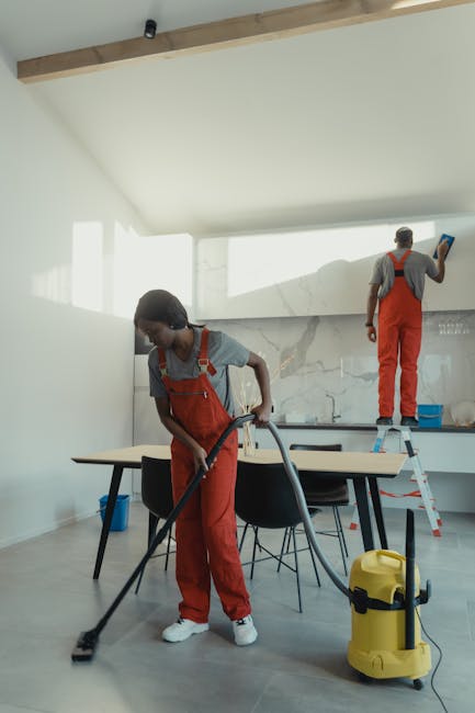 A professional cleaner engaging in domestic cleaning in a bright room with large windows and sheer curtains. The cleaner, wearing a black and white uniform, holds a plastic container filled with clean