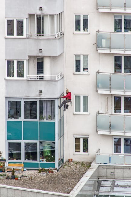 A professional window cleaner from Kennington Cleaners is ascending the exterior of a modern glass-fronted high-rise building using a harness and ropes for safety. The cleaner, wearing a helmet and sa