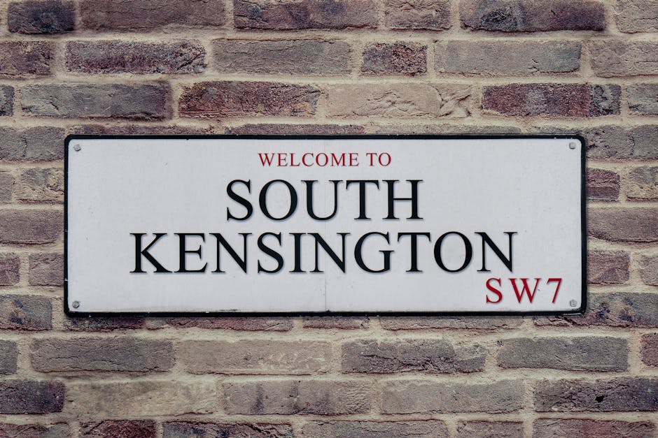 A rectangular white metal street sign mounted on a red brick wall, displaying the text 'Welcome to South Kensington SW7' in bold black and red lettering. The sign features four screw holes at each cor