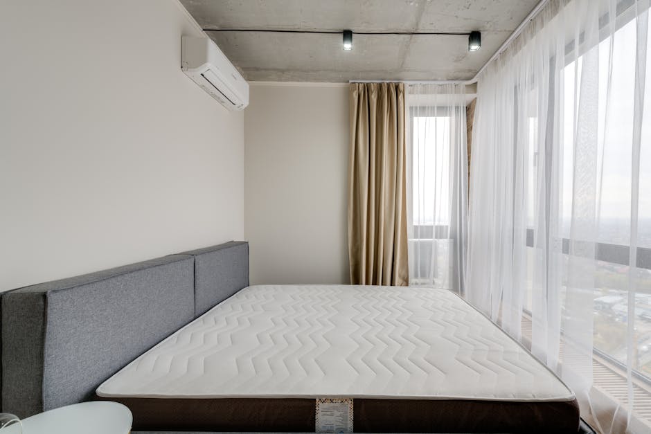 A neatly made bed with a dark upholstered headboard and frame, situated in a modern bedroom with dark green walls. The bed features a white fitted sheet, a neatly folded black throw or blanket at the