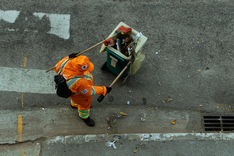 A person wearing white protective coveralls, safety goggles, and a respirator mask holding a yellow hazardous waste container outdoors on a landfill site filled with piles of assorted waste and debris