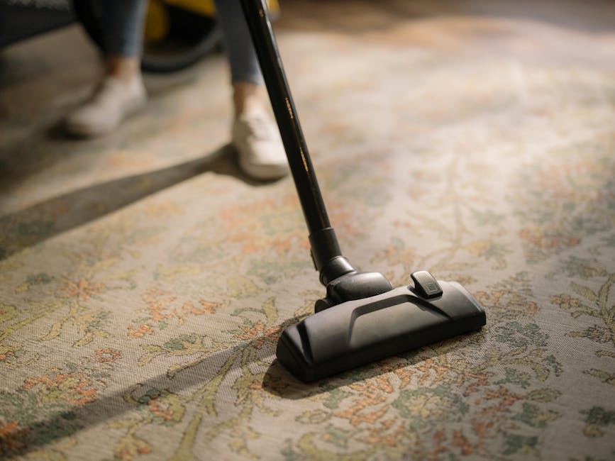 A close-up view of a black vacuum cleaner head actively cleaning a traditional patterned rug with floral motifs in shades of beige, green, and orange. The vacuum's hose and handle extend upward, with 